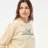 Buzo de cuello redondo y manga larga, confeccionado en felpa ligera color crema. Presenta un estampado frontal con la frase 'Still Dreaming' en color verde oscuro.