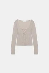 Chaqueta de punto color beige con escote en pico y manga larga, que incluye un top interior integrado con detalle de encaje a tono. Cierre frontal mediante botones.