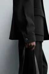 Chaquetón de corte regular fit en color negro, con cuello solapa, manga larga y cierre frontal con botones. Presenta dos bolsillos delanteros de plastrón con solapa en la cadera.