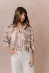 Camisa de rayas verticales blancas y marrones, de calce relajado y largo corto con volado en el ruedo. Presenta cuello camisero clásico, mangas tres cuartos con puños amplios, abotonadura frontal y un bolsillo aplicado en el frente.