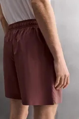 Pantalones cortos deportivos color marrón rojizo, confeccionados en tejido técnico ligero y elástico. Presentan cintura elástica ajustable con cordón interior, bolsillos frontales y un bolsillo trasero con cierre de cremallera. Los laterales tienen perforaciones para transpirabilidad y aberturas laterales en el bajo.