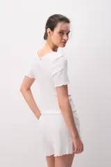 Remera de algodón blanca de manga corta, con diseño texturado, escote redondo y pequeño lazo decorativo en el centro. Presenta terminaciones con ondas en cuello, mangas y ruedo.