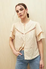 Camisa de algodón color beige con cuello mao, cierre frontal de botones y mangas cortas con volados. Presenta un diseño texturizado con rayas verticales y detalles de pasamanería calada en el frente.