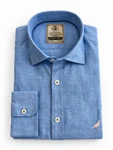 Camisa de lino color celeste con cuello corto (short collar), corte clásico, manga larga y botones frontales blancos con costura cruzada. Cuenta con un bordado sutil de un pájaro en el sector inferior.