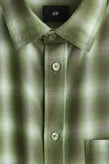 Camisa de corte relajado confeccionada en mezcla de algodón suave, con estampado a cuadros en tonos verdes y blancos. Presenta cuello inglés, bolsillo frontal en el pecho, cierre de botones y mangas largas con puños ajustables.