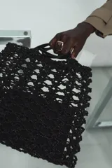 Bolso tipo shopper pequeño de crochet negro, con asas de hombro de material liso negro y una bolsa interior con cierre de cremallera.