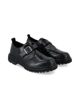 Zapato tipo monk strap de cuero negro con textura efecto cocodrilo, suela track dentada y cierre con hebilla metálica.