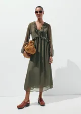 Vestido midi color verde militar, confeccionado en tejido fluido de poliéster. Presenta un diseño evasé, escote de pico con volantes, y mangas largas con puños elásticos. Incluye detalles de volantes en el cuello y forro interior.