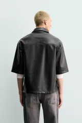 Sobrecamisa de corte relajado confeccionada en tejido efecto piel, con cuello solapa, manga corta y cierre frontal con cremallera. Presenta un bolsillo plastrón en el pecho y bajo con aberturas laterales.