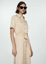 Vestido midi de tejido satinado color beige, con cuello camisero, cierre frontal de botones y manga corta. Presenta diseño recto con lazo ajustable en la cintura y dos bolsillos de parche en el pecho.