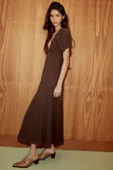 Vestido midi de color marrón, confeccionado en tejido de gasa con textura. Presenta un escote en pico con detalle de lazada frontal, mangas cortas abullonadas con terminación elástica y falda con detalle de godets que aportan volumen. Incluye forro interior a tono y cierre lateral con cremallera oculta.