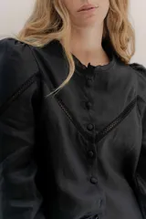 Camisa de lino color beige con mangas abullonadas y detalles de puntilla negra en forma de V en el frente. Presenta cuello a la base con borde desflecado, cierre frontal con botones forrados y puños con alforzas.