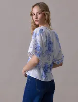 Blusa blanca estampada con flores azules, escote en V y mangas cortas amplias. Detalles de puntilla en escote y mangas.