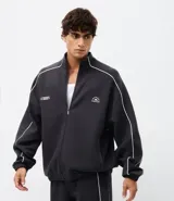 Buzo deportivo de corte boxy, color negro, con cuello alto, cierre frontal completo y mangas largas. Presenta detalles de líneas blancas a lo largo de las mangas y lettering estampado en el pecho.