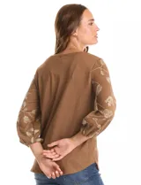 Blusa de tejido fino color marrón con bordado floral en hilo dorado. Presenta cuello redondo, mangas tres cuartos abullonadas con puños elásticos fruncidos y ruedo redondeado con corte asimétrico.