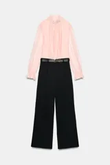 Mono largo con parte superior de blusa rosa de manga larga, cuello alto fruncido y puños con volantes. La parte inferior es un pantalón negro de pernera ancha con bolsillos delanteros y un cinturón negro con hebilla dorada.