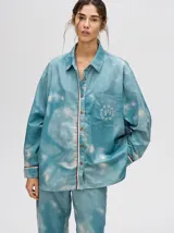 Camisa oversize de viscosa con estampado tipo tie-dye en tonos azulados. Presenta cuello clásico, cierre frontal con botones, bolsillo frontal con bordado y detalles de vivos en contraste en la tapeta y puños.