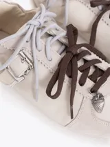 Zapatillas de cuero vacuno en color beige, con recortes de gamuza en la puntera y talón, doble cordón en tonos gris y marrón, hebilla lateral y dije colgante en la cordonera. Suela plana de goma.
