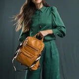 Mochila de símil cuero color marrón con apertura superior y tres bolsillos frontales con cierres metálicos. Cuenta con bolsillos laterales y una correa de galón con diseño geométrico, regulable y extraíble, que permite su uso como bandolera.