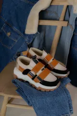 Zapatillas deportivas color beige con detalles en negro y marrón. Presentan una suela gruesa con diseño de tractor y ajuste con tiras de velcro color caramelo.