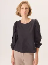 Camisa negra con cuello redondo, mangas largas con puños elásticos y detalle de volados en los hombros. Presenta un diseño combinado con frente y mangas de tela de camisa y espalda de tela de remera.