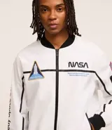 Campera tipo bomber blanca con detalles en negro, inspirada en uniformes espaciales de la NASA. Presenta parches con el logo de la NASA, la bandera de EE. UU. y texto en la manga que dice "Kennedy Space Center".