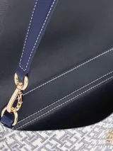 Cartera tipo mensajera con solapa, confeccionada en poliuretano con estampado integral del monograma TH. Presenta detalles en color azul marino, herrajes dorados y el logo metálico TH en el frente. Incluye asa superior extraíble y cierre a presión.