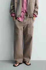 Pantalón de corte relajado en color marrón con efecto lavado. Presenta bolsillos frontales tipo cargo, cierre con cremallera y botón, y una línea horizontal de tachas metálicas a tono en la parte superior de las piernas.