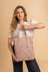 Bolso tipo tote bag de símil cuero color rosa pálido, con asas dobles y detalles de costura en color beige claro. Presenta un bolsillo frontal rectangular aplicado y herrajes metálicos.