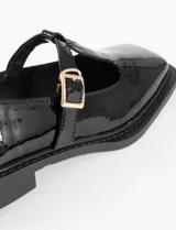 Mocasín estilo T-bar color negro en charol, con punta cuadrada, tira en el empeine con hebilla dorada y suela plana.