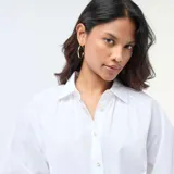 Camisa de popelina blanca con cuello clásico, cierre frontal de botones y mangas largas abullonadas con puños ajustados.