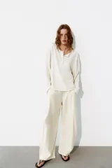 Pantalón fluido de color blanco, de tiro alto con trabillas y pliegues en el delantero. Cuenta con bolsillos laterales, cierre frontal con cremallera, botón interior y gancho metálico.