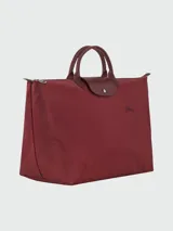 Bolso de viaje de gran tamaño color verde, confeccionado en lona con detalles de cuero. Presenta asas superiores cortas, solapa con broche de presión y cierre de cremallera. Su diseño es ligero y plegable.