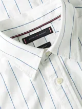 Camisa de manga larga de Tommy Hilfiger, color blanco con finas rayas verticales celestes, corte amplio, cuello clásico, cierre frontal con botones, un bolsillo de parche en el pecho con el logo bordado y puños abotonados.