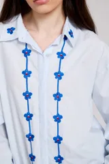 Camisa blanca de corte holgado con cuello camisero y cierre frontal de botones. Presenta un delicado bordado vertical de flores pequeñas en color bordó a lo largo de la botonera y en el cuello.