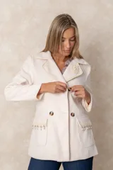 Blazer cruzado color beige, con cierre de cuatro botones frontales. Presenta detalles artesanales de encaje con perlas y cristales en la solapa y en los bolsillos delanteros.