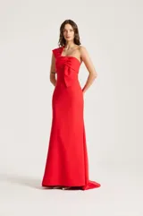 Vestido largo de fiesta color rojo, de un solo hombro, con detalle de lazo drapeado en el pecho y silueta ajustada que se ensancha levemente hacia el suelo con una sutil cola trasera.