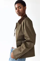 Campera de sarga color verde militar, con cuello camisero de pana marrón oscuro, cierre frontal con solapa y botones a presión metálicos, bolsillos frontales grandes con solapa y mangas raglán.