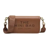 Cartera bandolera Marc Jacobs modelo The Mini Bag, confeccionada en cuero premium color beige. Presenta diseño rectangular con solapa frontal, logo grabado en el frente y correa ajustable con el nombre de la marca tejido.