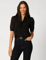 Blusa negra de corte clásico con escote en V y lazo integrado al cuello. Presenta mangas cortas ligeramente abullonadas confeccionadas en tejido semitransparente con puños ajustados.
