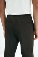 Pantalón tipo jogger de vestir color marrón oscuro, con cintura elástica y cordón ajustable. Presenta pliegues marcados en la parte delantera y un corte slim fit.