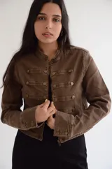 Chaqueta de estilo militar en color marrón, con corte estructurado y cuello mao. Presenta cierre frontal con cremallera y detalles de filas horizontales con botones metálicos decorativos en el frente y en los puños.