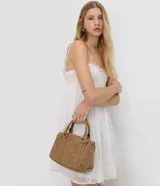 Cartera satchel de gamuza sintética color marrón, con diseño estructurado, doble asa de mano y tachas decorativas en los bordes. Cuenta con cierre superior y herrajes plateados.