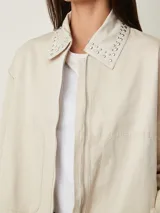Campera cruda de gabardina con cierre frontal, bolsillos con cierre y cuello mao adornado con tachas plateadas.