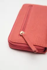 Billetera de formato rectangular con cierre perimetral, confeccionada en material símil cuero de textura suave. Presenta un aplique metálico circular con logo grabado en el frente y tirador de cierre en el mismo material.