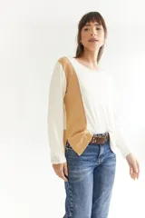 Remera de manga larga con diseño bicolor, combinando paneles en color crudo y tostado. Presenta un corte asimétrico con el frente más corto que la espalda, aberturas laterales en el ruedo y costuras decorativas a la vista.