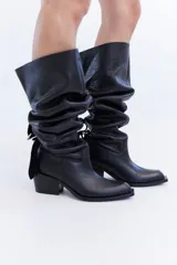 Bota de cuero negro de caña alta slouchy, con diseño fruncido ajustable mediante un juego de correas y hebillas. Presenta taco bajo cuadrado y punta cuadrada.