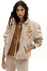 Chaqueta oversized de gamuza de cabra color arena, con apliques tribales en cuero de oveja color mostaza. Presenta hombros exagerados, cuello camisero redondeado, bolsillos frontales y botones a presión forrados. Cuenta con forro capitoneado y un bolsillo interno.