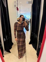 Vestido largo de corte ajustado, con cuello redondo y mangas largas. Presenta un diseño de estampado a cuadros en tonos marrones y un detalle de fruncido lateral en la cintura.
