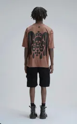 Remera manga corta color marrón (mocca) con un gran estampado trasero en negro que presenta tres máscaras o calaveras estilizadas con una corona en la parte superior y gotas o lágrimas cayendo.